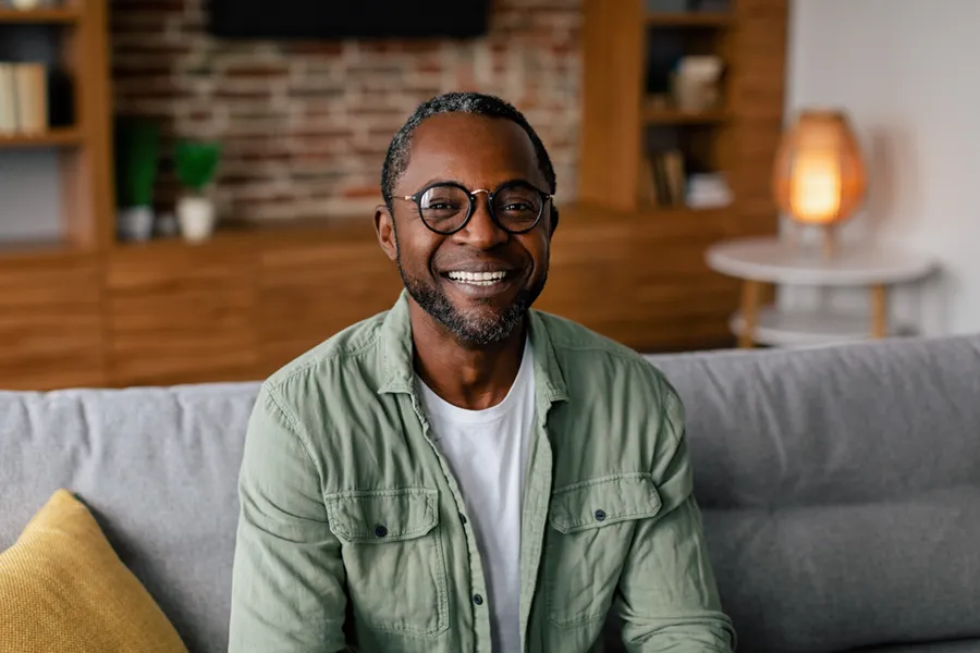 Andropause-Doctor A middle aged black man in a green button up shirt sits on his couch smiling, happy with his treatment for andropause and low testosterone from Blessed Beauty Boutique in Greater Phoenix.