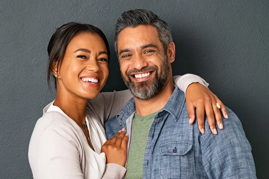 A middle-aged couple with their arms around each other standing against a gray background and smiling, happy with the bioidentical hormone therapy they received from Blessed Beauty Boutique in Greater Phoenix.