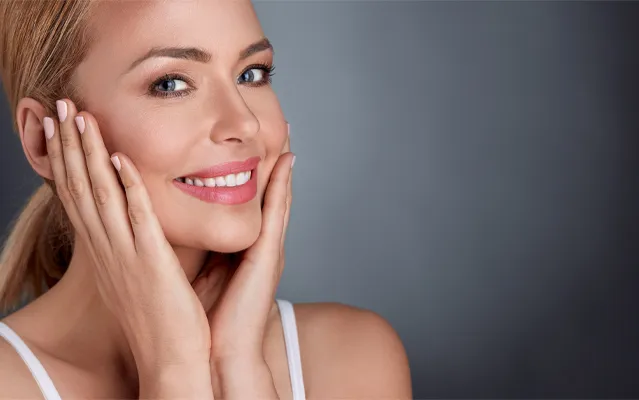 Facial-Clinic A middle-aged blonde woman with beautiful skin smiles and holds her face. Get facial treatments from Blessed Beauty Boutique in Greater Phoenix.