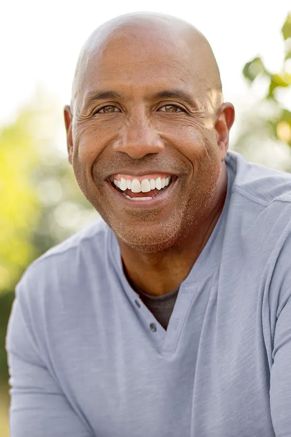 Healthy-Aging-Peptides-Treatment A middle-aged black man in a gray v-neck t-shirt sits outside smiling, happy with the healthy aging peptide treatments from Blessed Beauty Boutique in Greater Phoenix