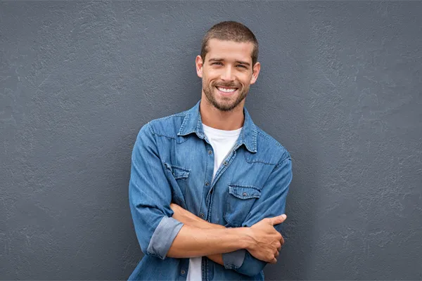 Hormones-for-Men-Doctor A man in a denim shirt stands smiling against a gray-blue wall, pleased with his testosterone hormone therapy from Blessed Beauty Boutique in Greater Phoenix.