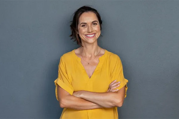 Hormones-for-Women-Clinic-2 A woman in a green shirt stands smiling against a gray-blue wall, pleased with her hormone therapy from Blessed Beauty Boutique in Greater Phoenix.