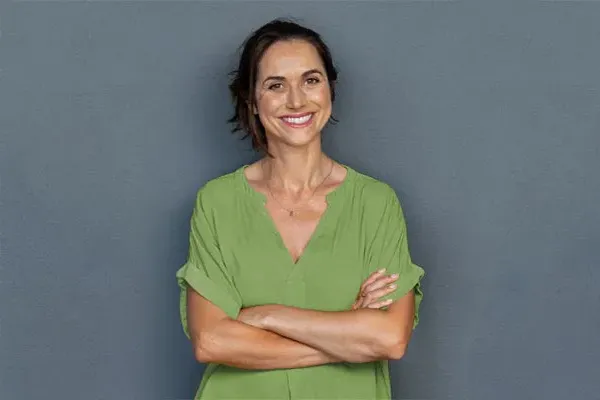 A woman in a green shirt stands smiling against a gray-blue wall, pleased with her hormone therapy from Blessed Beauty Boutique in Greater Phoenix.
