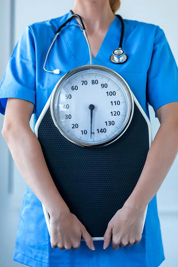 Medical-Weight-Loss-Treatment A nurse in blue scrubs with a stethoscope hanging around her neck holds a scale, representing weight loss treatments from Blessed Beauty Boutique in Greater Phoenix.