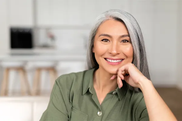 Menopause-Clinic An asian woman with long gray hair and a green shirt sitting and smiling on her couch, happy with her menopause care from [NAME} in Greater Phoenix.