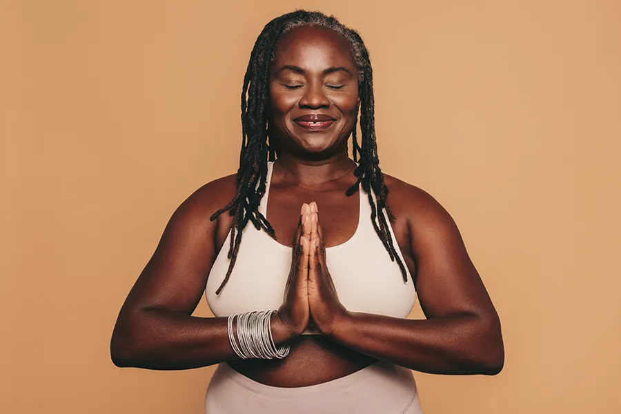 Obesity-Clinic An older African American woman confidently posing against a warm background smiling with hands in a prayer pose. Blessed Beauty Boutique treats obesity using functional medicine in Greater Phoenix.
