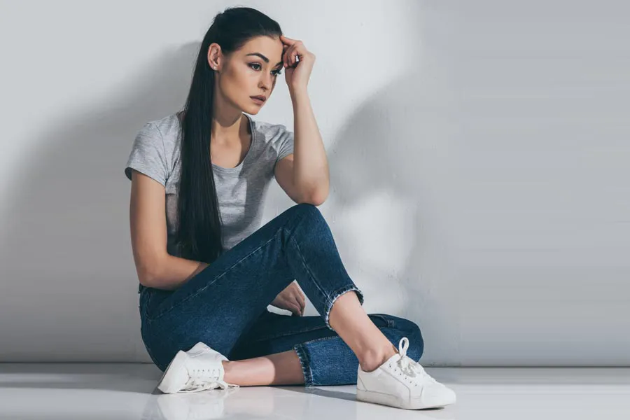 PCOS-Doctor A young brunette woman in a gray t-shirt and jeans sits against a gray wall with her head resting on her hand and a distressed expression. Get help for Polycystic Ovarian Syndrome (PCOS) from Blessed Beauty Boutique in Greater Phoenix.