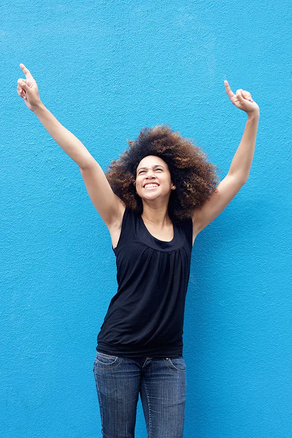 PMS-Treatment A woman in a dark blue tank top standing in front of a bright blue wall, raising her arms in celebration of relief from PMS from Blessed Beauty Boutique in Greater Phoenix.