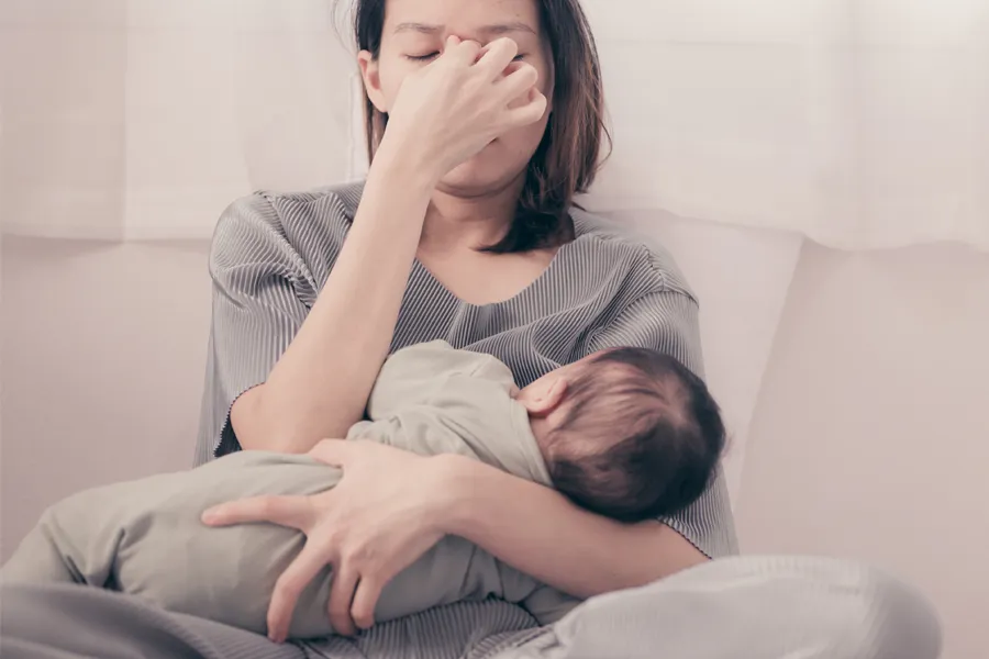 Postpartum-Hormone-Imbalance-Clinic A woman nursing an infant on couch rubs her eyes with exhaustion. Get treatment for postpartum hormone imbalance from Blessed Beauty Boutique in Greater Phoenix.