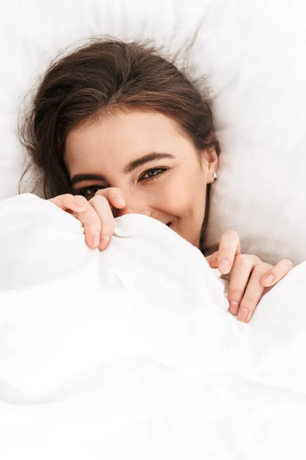 Vaginal-Rejuvenation-and-Vulvar-Wellness-Treatment A woman in bed holds the covers over her mouth playfully. Get Vaginal Rejuvenation and Vulvar Wellness treatment from Blessed Beauty Boutique in Greater Phoenix.